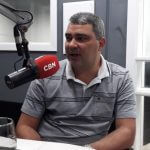 Imagem de um estúdio de rádio, com o Dr João Alberto Prust, usando uma camisa cinza, está sentado em frente à um microfone, sendo entrevistado pela rádio CBN.