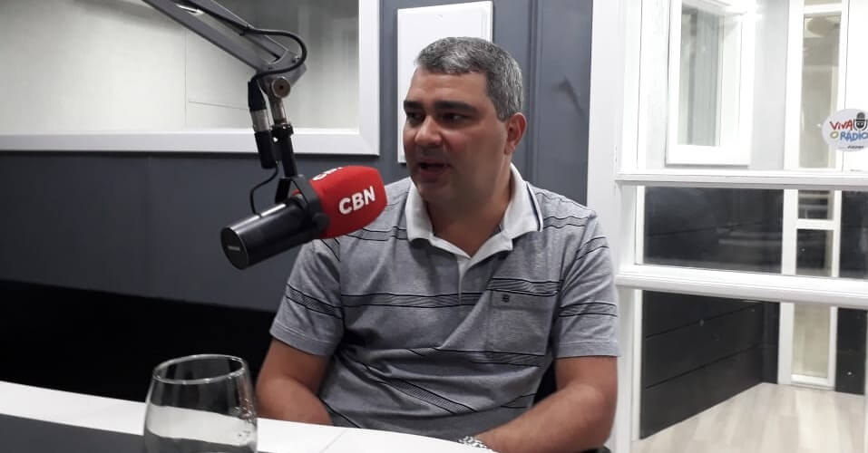 Imagem de um estúdio de rádio, com o Dr João Alberto Prust, usando uma camisa cinza, está sentado em frente à um microfone, sendo entrevistado pela rádio CBN.